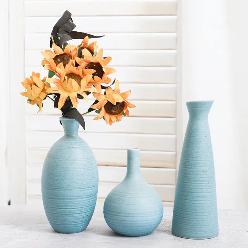 Three light blue vases of varying heights, with a bouquet of orange sunflowers in the tallest vase, set against a white background.