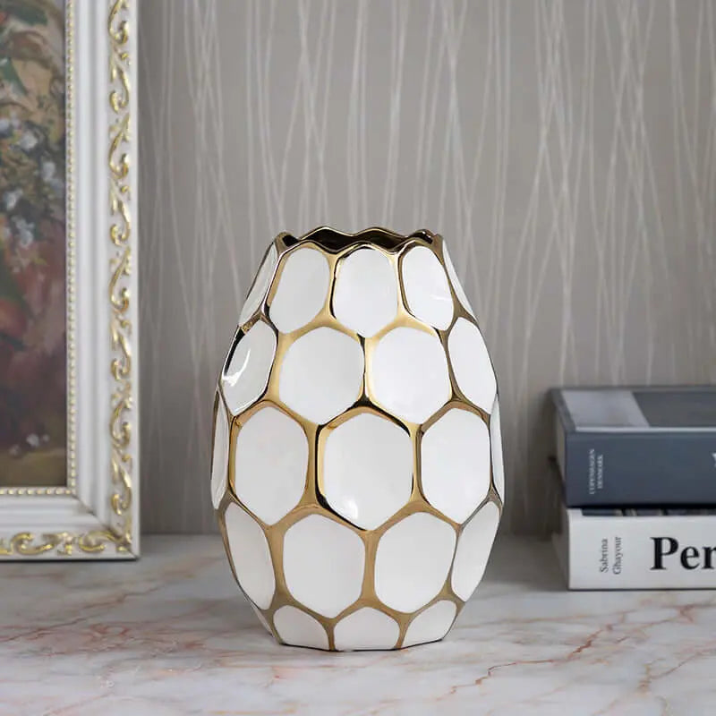 A decorative white vase with a honeycomb pattern and gold accents, placed on a marble surface near a framed painting and books.