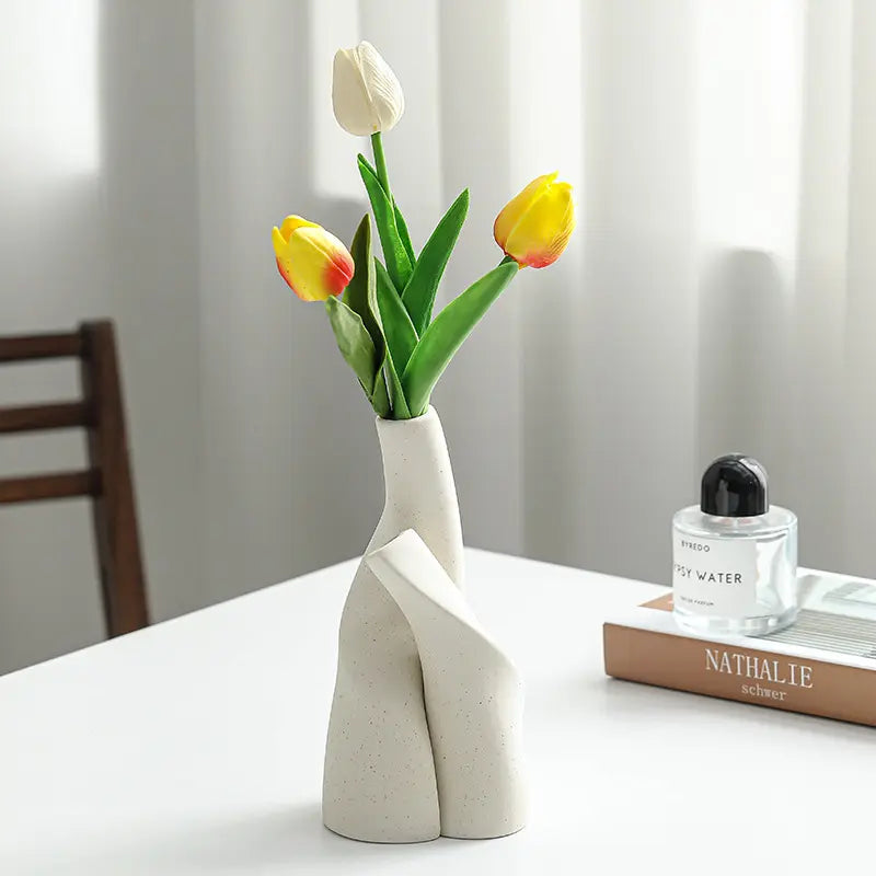 A modern white vase holding vibrant yellow and white tulips sits on a table beside a perfume bottle and a book.