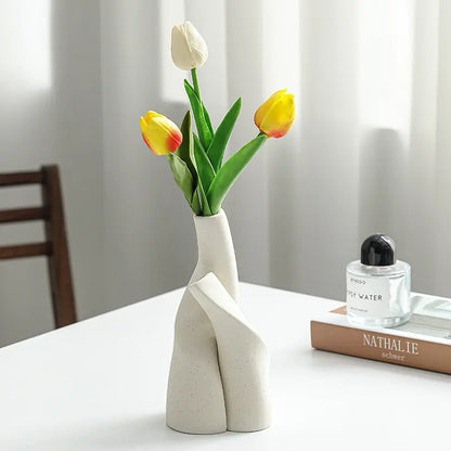 A modern white vase holding vibrant yellow and white tulips sits on a table beside a perfume bottle and a book.