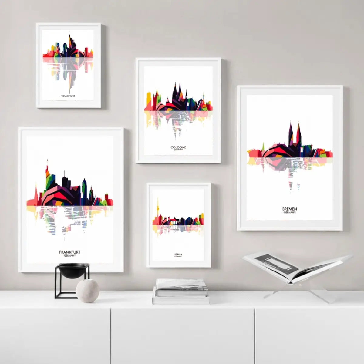 A collection of colorful city skyline art prints featuring Frankfurt, Cologne, Bremen, and Berlin, displayed in white frames on a wall.