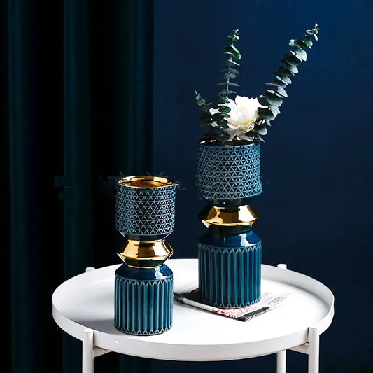 Two decorative vases in blue and gold, one with a flower and eucalyptus branches, placed on a white side table against a dark backdrop.