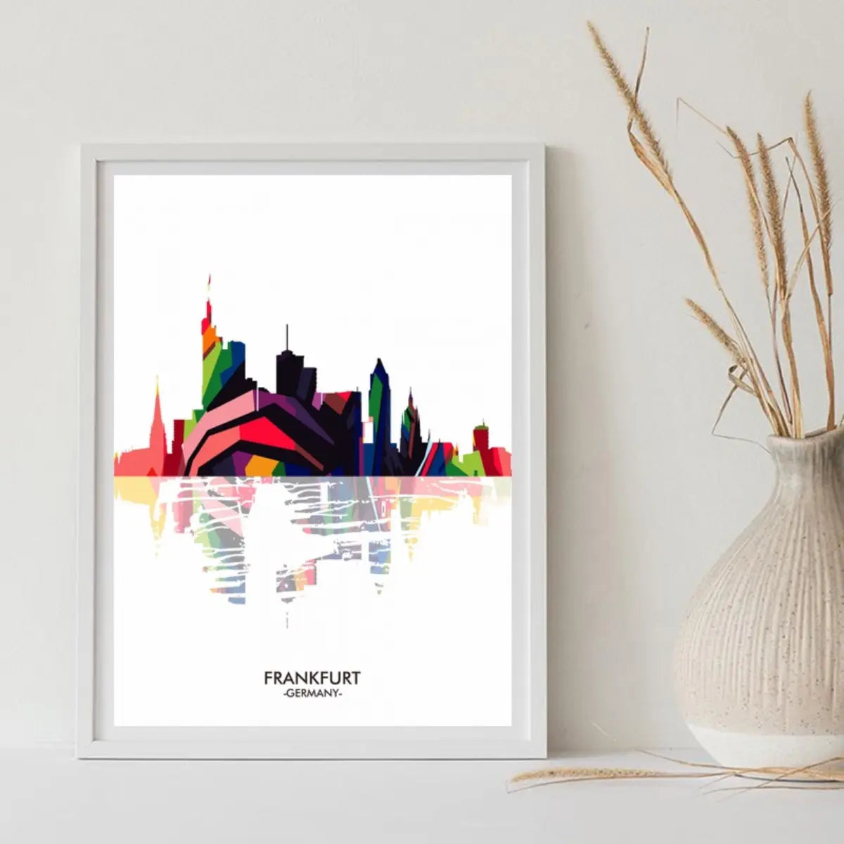 Colorful silhouette of Frankfurt skyline with reflections, displayed in a white frame, alongside a decorative vase and dried plants.