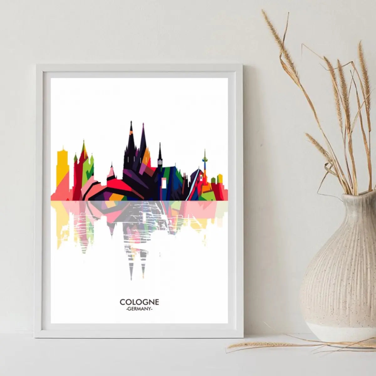 A colorful abstract skyline of Cologne, Germany, displayed in a white frame, with reflections below.