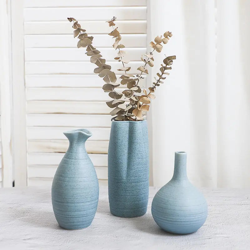 Three textured blue vases of varying shapes, one holding dried eucalyptus stems, against a light backdrop.