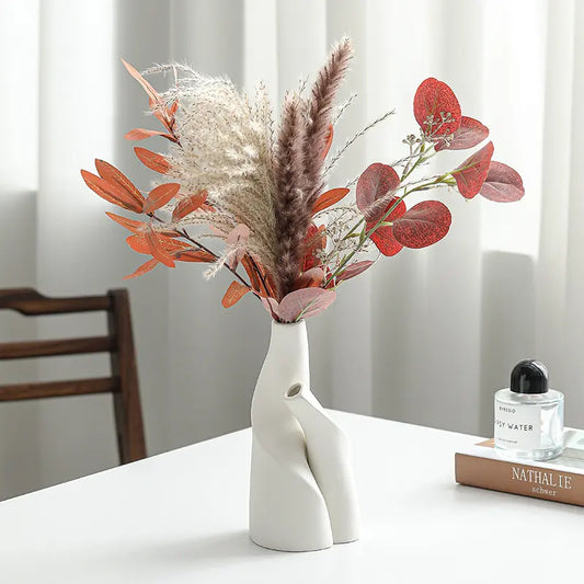A white hand-shaped vase holds an arrangement of dried flowers and foliage in warm autumn tones, next to a perfume and books on a table.