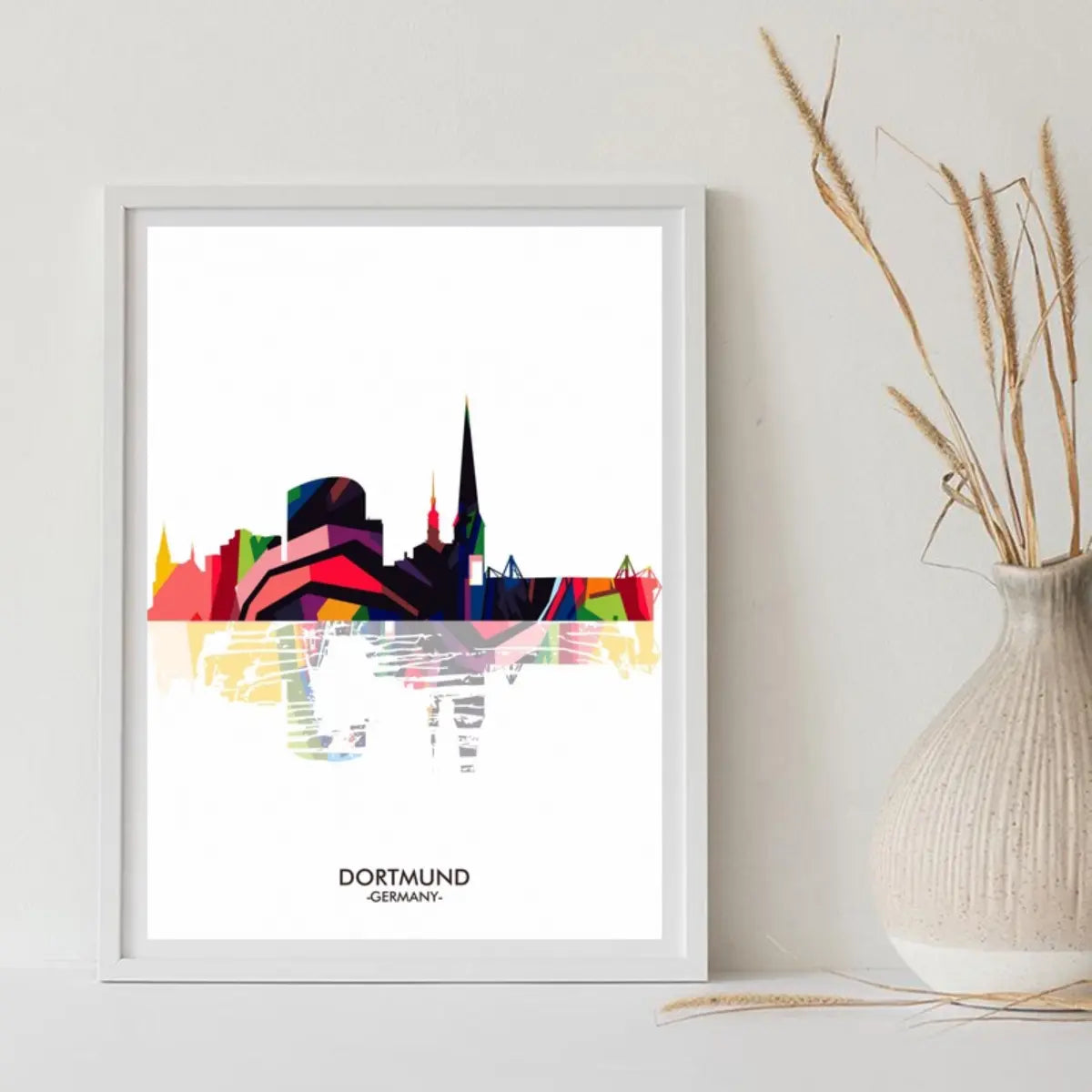 Colorful abstract skyline of Dortmund, Germany, displayed in a white frame beside a textured vase and dried grasses.