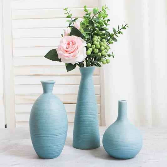 Three blue ceramic vases of varying shapes, one with pink roses and greenery, set against a white backdrop.