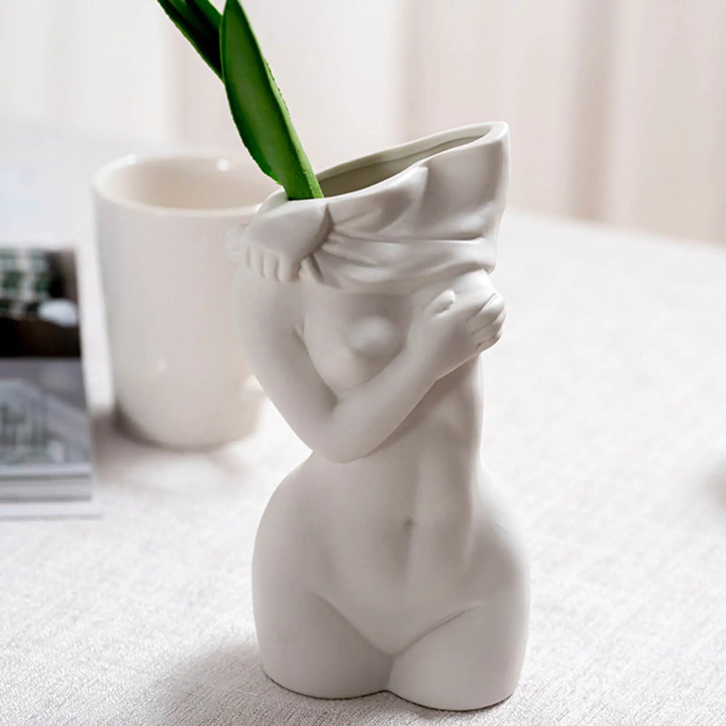 A white ceramic vase shaped like a nude female figure, holding a green plant, set on a light fabric surface.