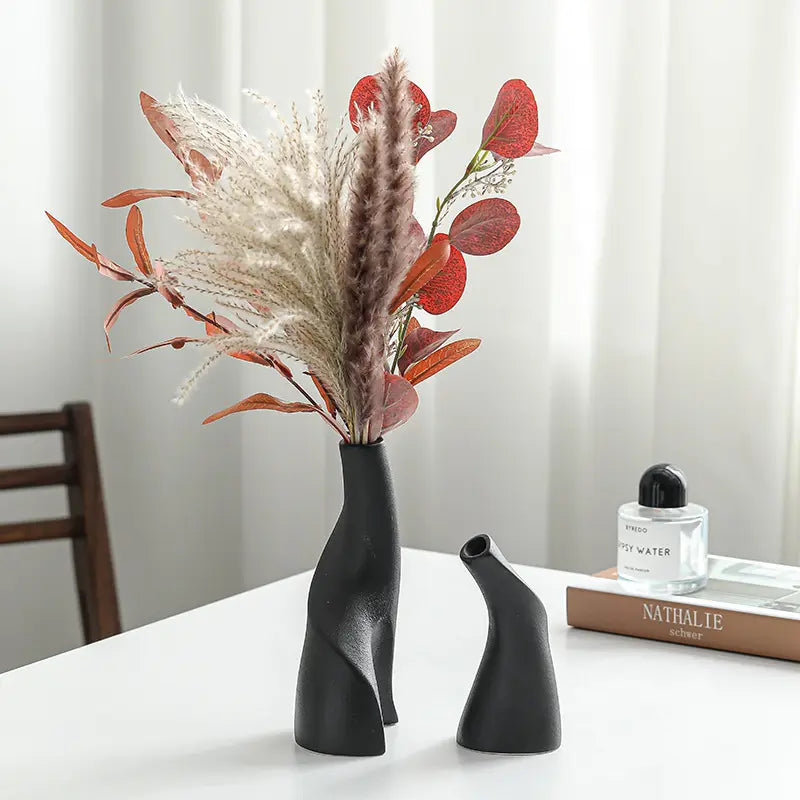 A stylish black vase filled with dried flowers and leaves, accompanied by a smaller vase, set against a light background.