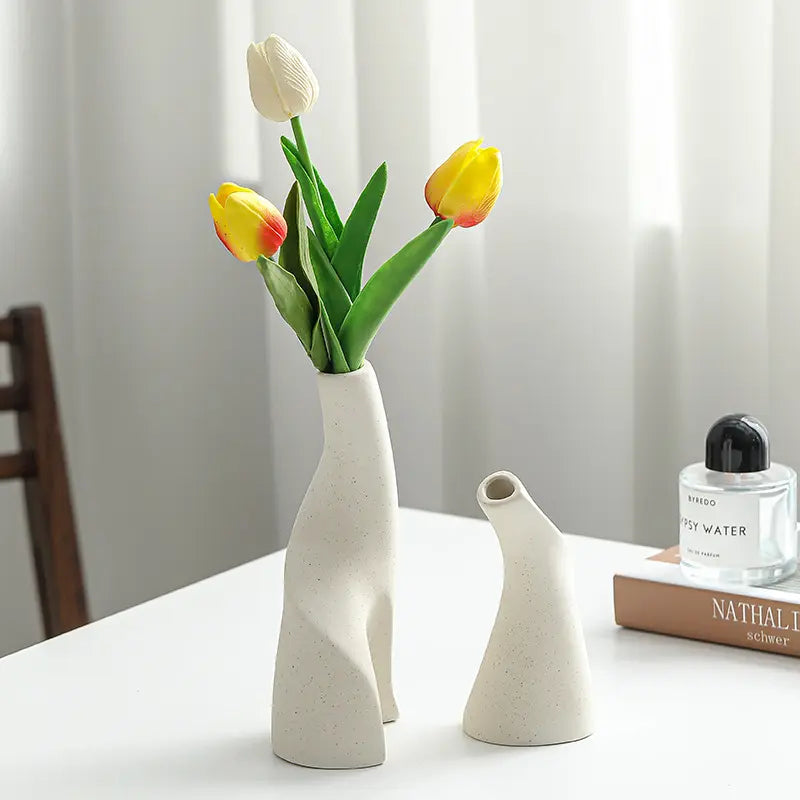 A modern beige ceramic vase holds colorful tulips against a light background, beside a small vase and a perfume bottle.