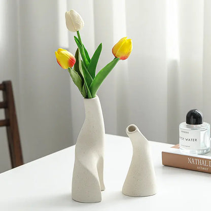 A modern beige ceramic vase holds colorful tulips against a light background, beside a small vase and a perfume bottle.