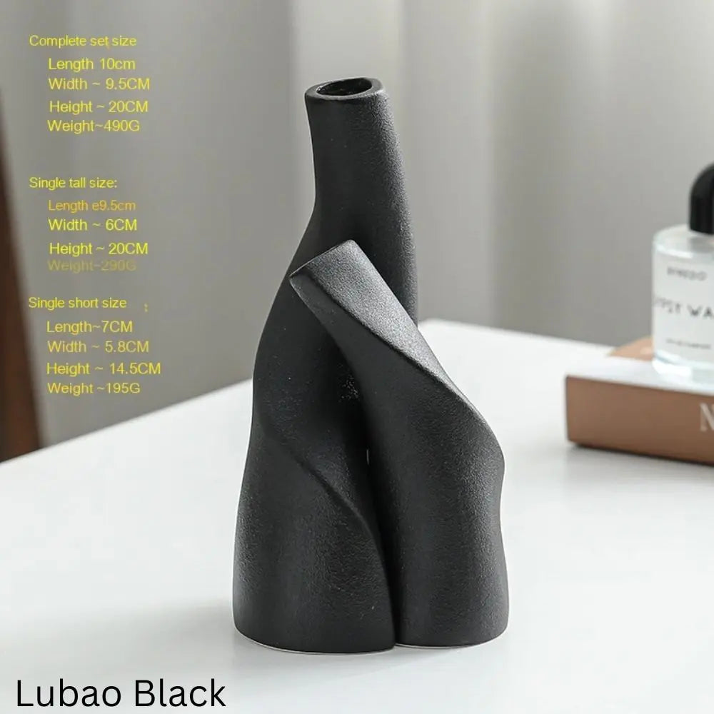 A modern black vase with a sleek, abstract design, positioned on a table with a nearby perfume bottle and books.